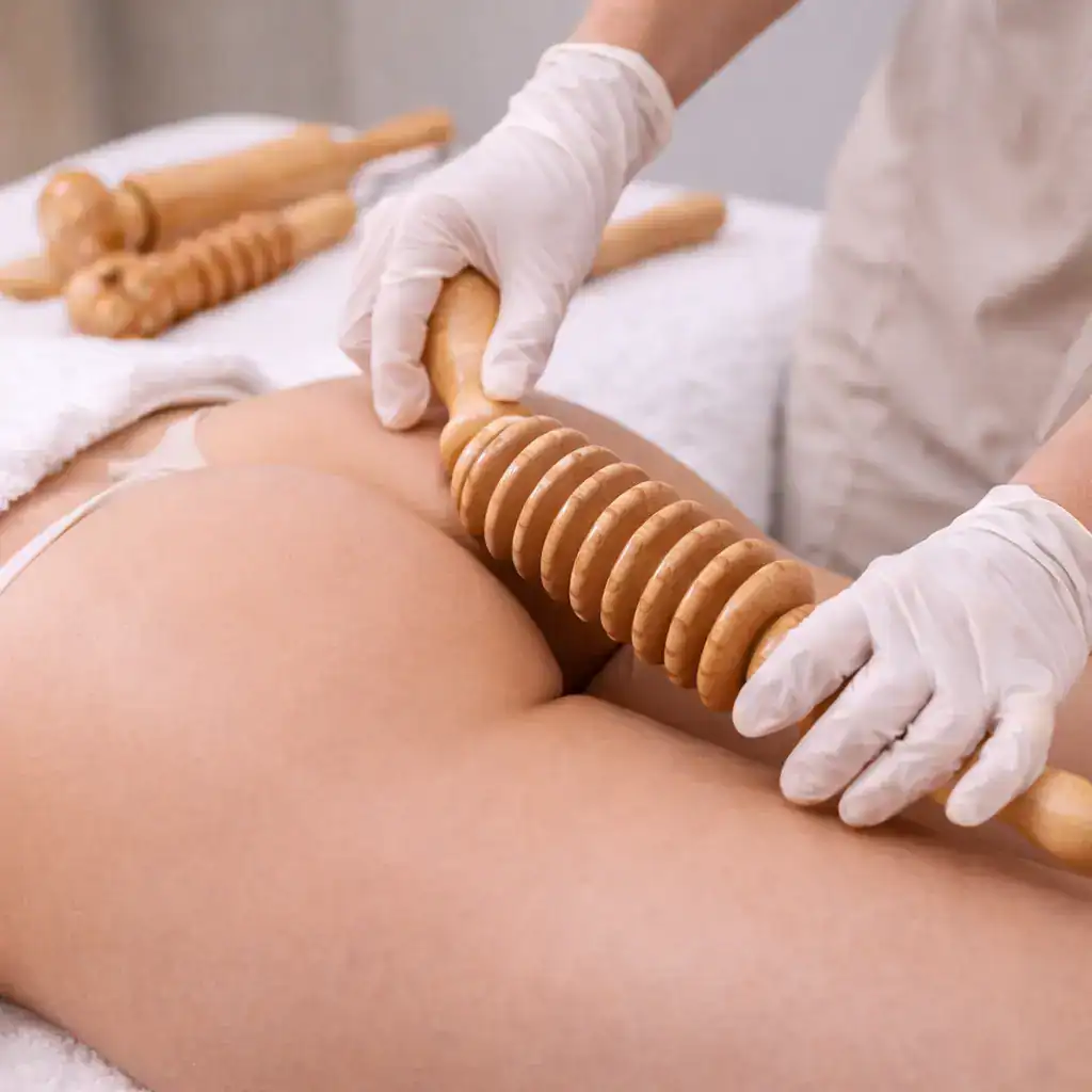 A gloved hand moving a ridged wooden roller along a woman's upper thigh, with more wooden tools on a towel